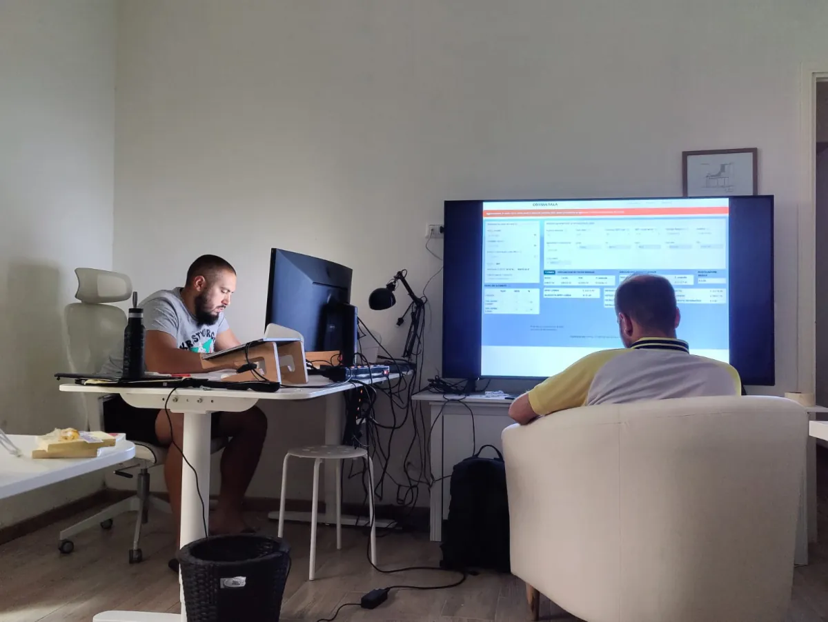 Two developers at their desks in a bright room, one facing a large wall-mounted monitor