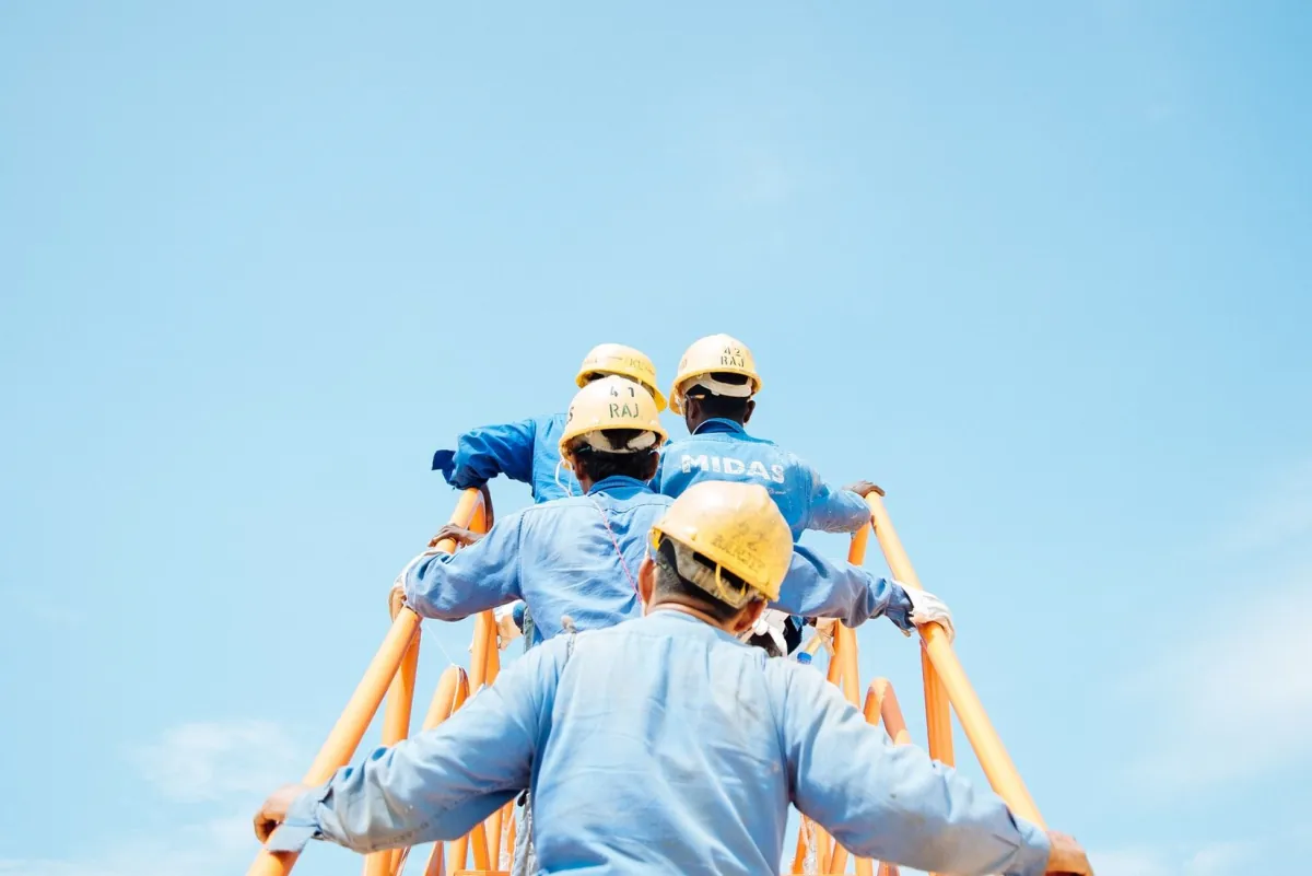 Workers in yellow helmets climbing a tall steel structure against a blue sky